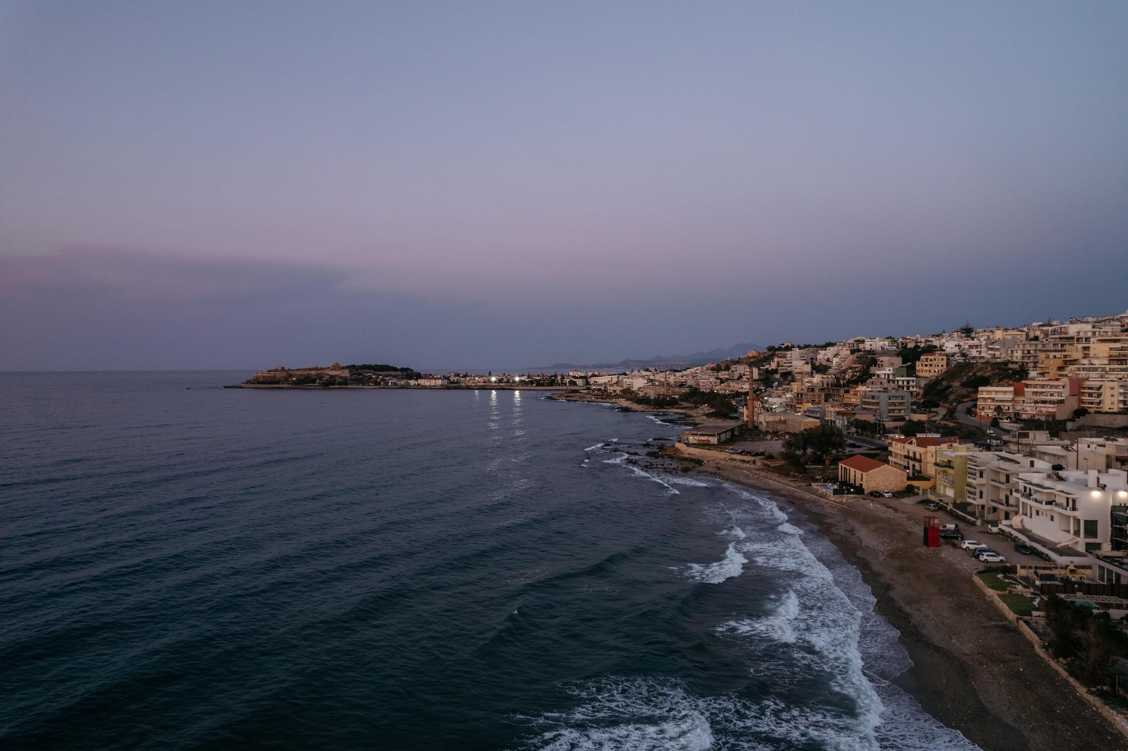 Une ville côtière avec des bâtiments qui bordent le littoral, une plage et une petite île visible au loin.