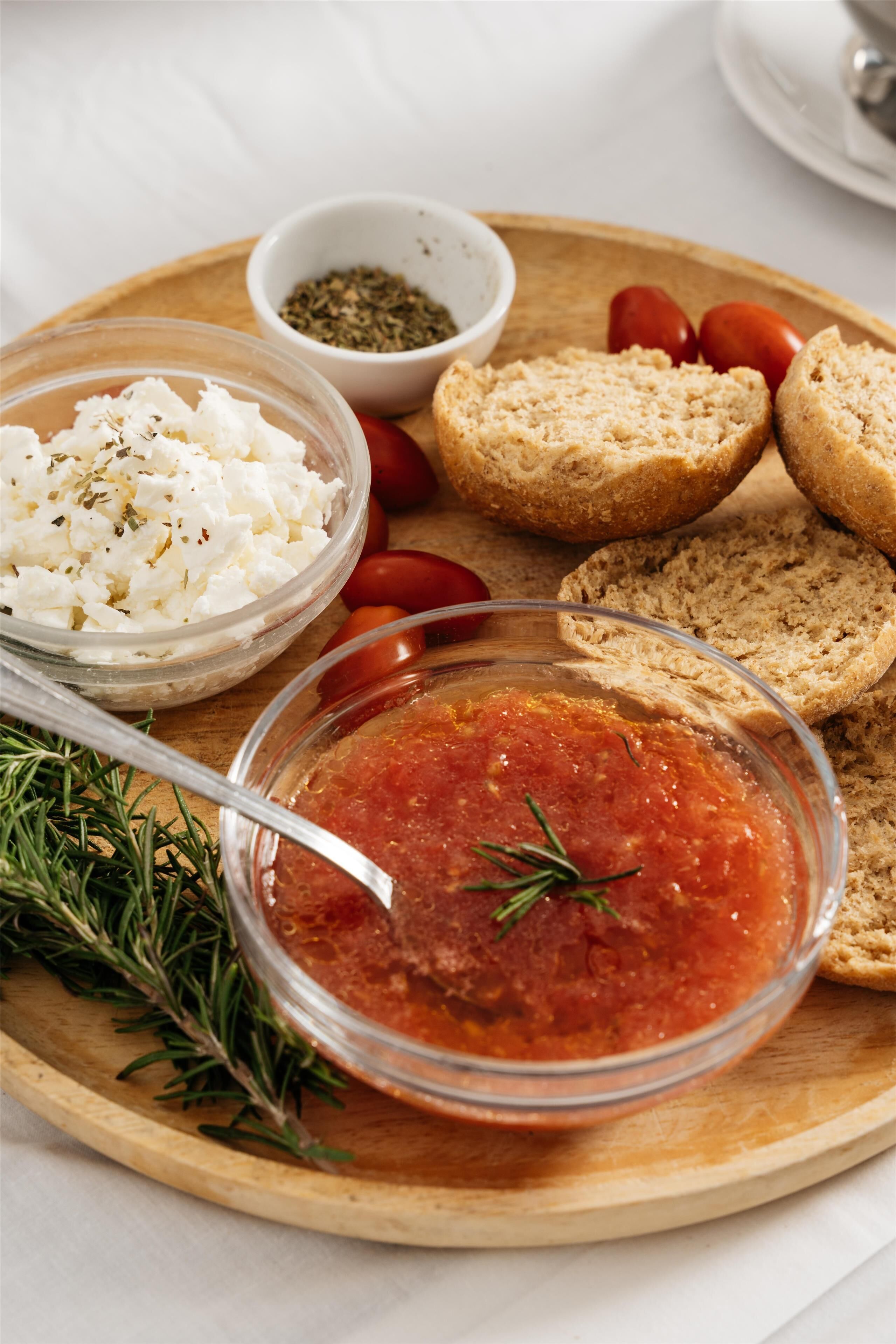 A wooden board with various food items including a bowl of white cheese, a bowl of red sauce, and some bread.