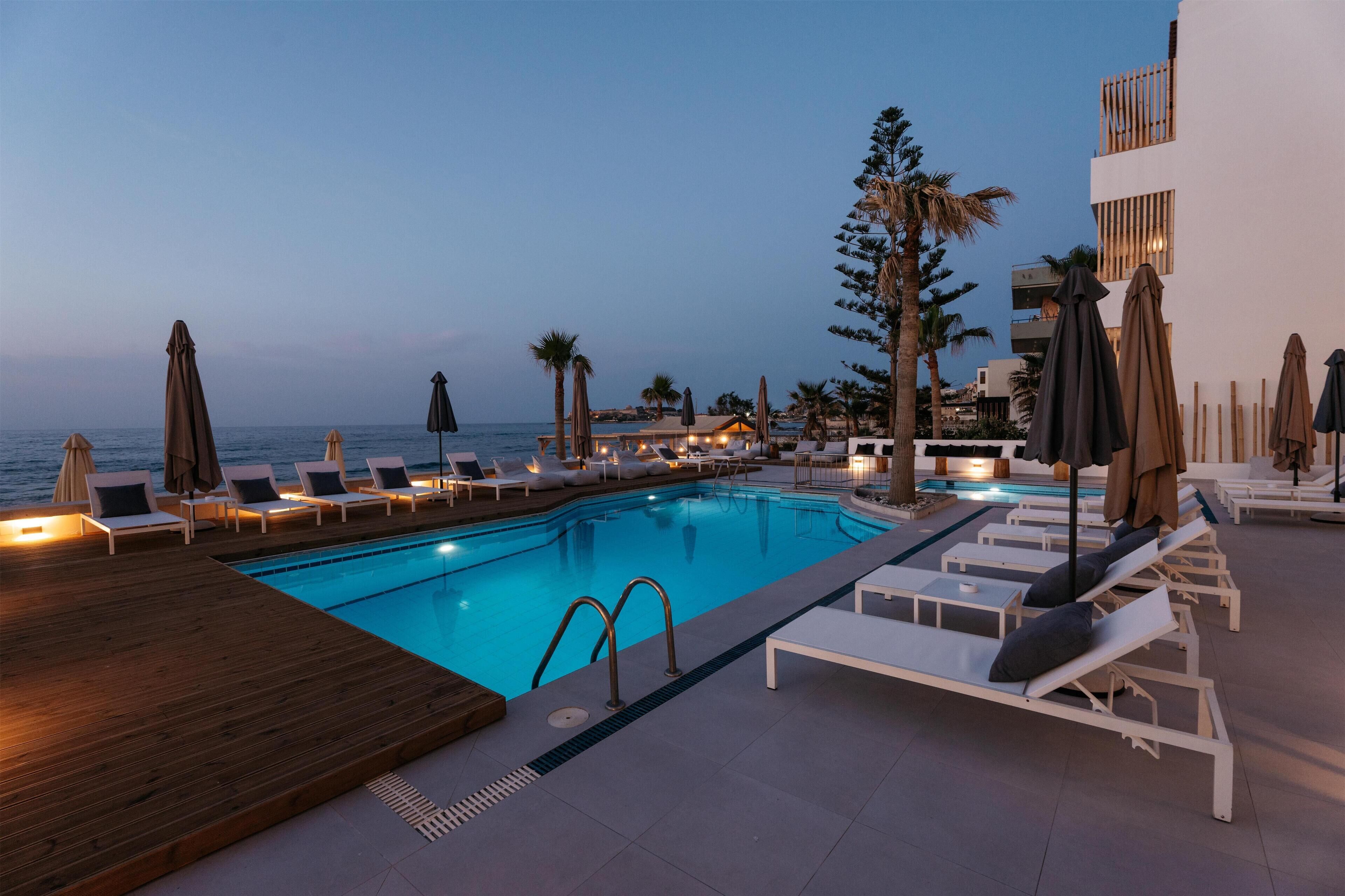 A poolside area with lounge chairs and umbrellas, a wooden deck, and a view of the sea.