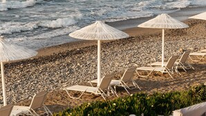 A beach with lounge chairs and umbrellas, a rocky shoreline, and a distant view of a fortified structure. - Petradi Beach Lounge Hotel by GHH (Rethymno)