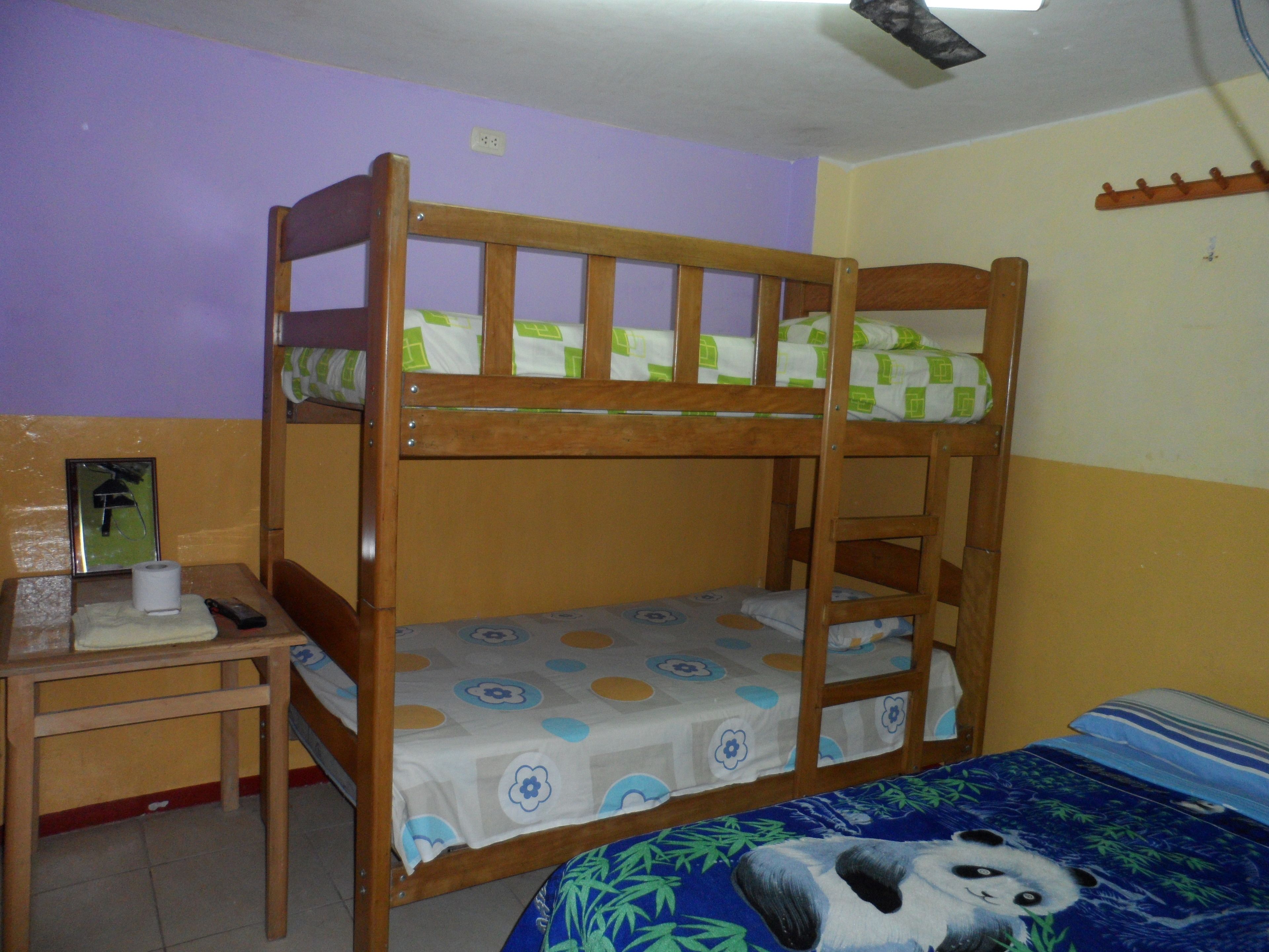 Una cama litera con estructura de madera, una mesita de noche con un rollo de papel higiénico, y una cama con un cubrecolcha de temática de panda.