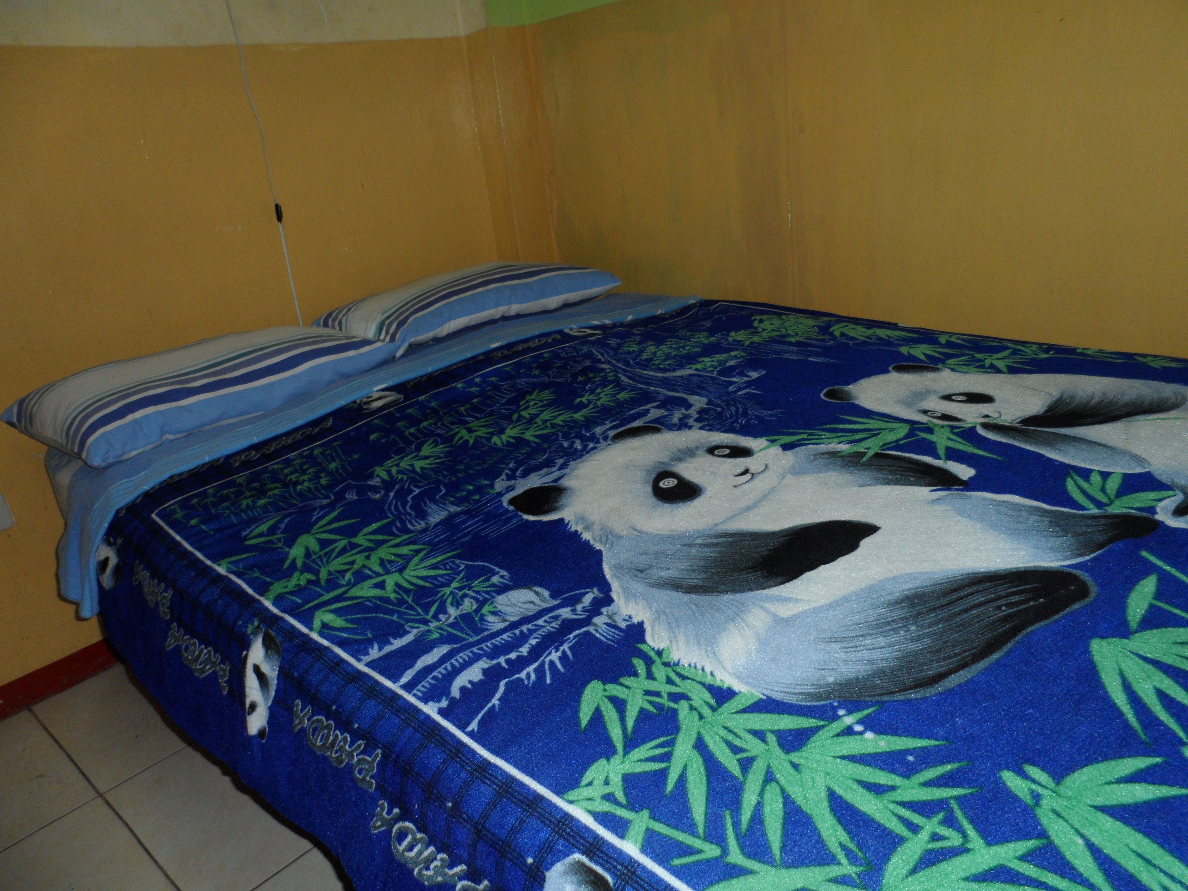 Una cama con cubrecama azul decorada con ilustraciones de pandas y hojas de bambú, junto a dos almohadas a rayas.