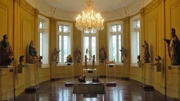 Un salón majestuoso con una gran araña, estatuas sobre pedestales y altos ventanales.