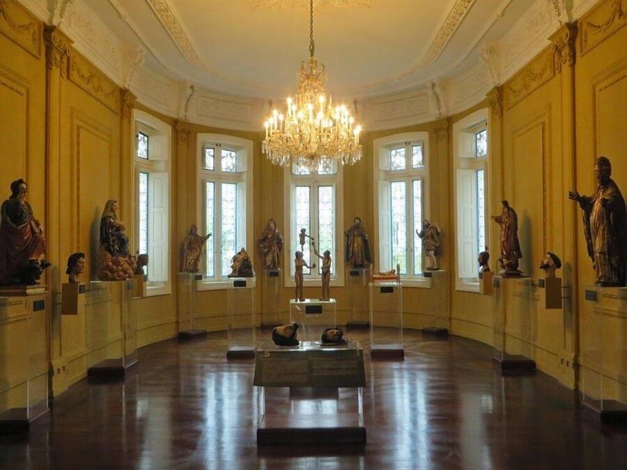 A grand hall with a large chandelier, statues on pedestals, and tall windows.
