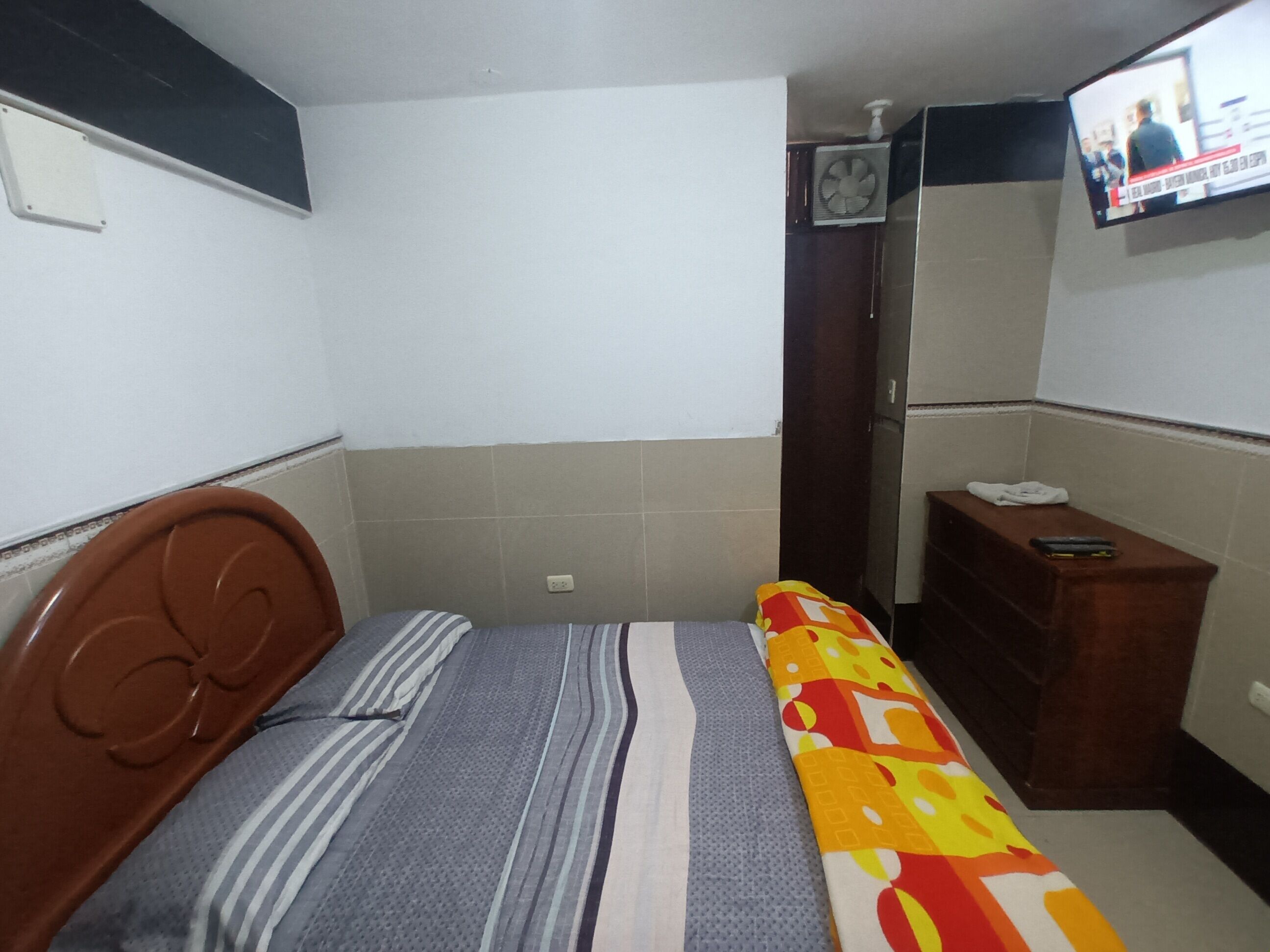 Una habitación pequeña y sencilla con una cama, un aparador de madera, un televisor y un ventilador.