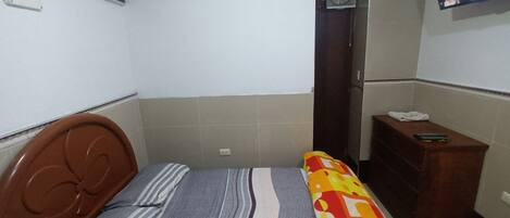 Una habitación pequeña y sencilla con una cama, un aparador de madera, un televisor y un ventilador.
