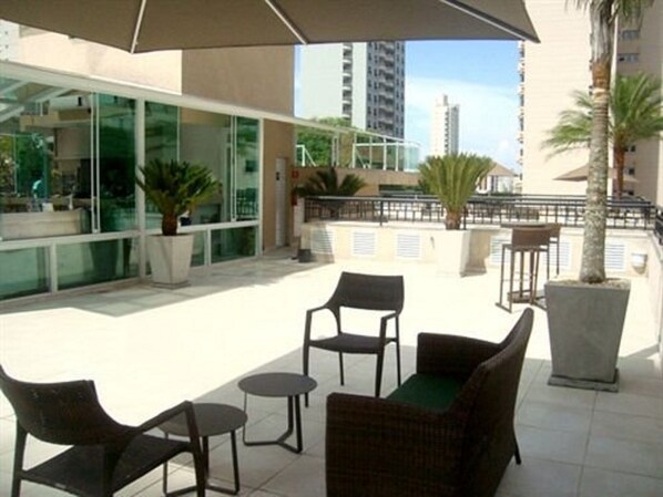 Un área de estar al aire libre con mobiliario de mimbre y un paraguas grande, con vistas a un edificio moderno con ventanas de vidrio y una palmera.