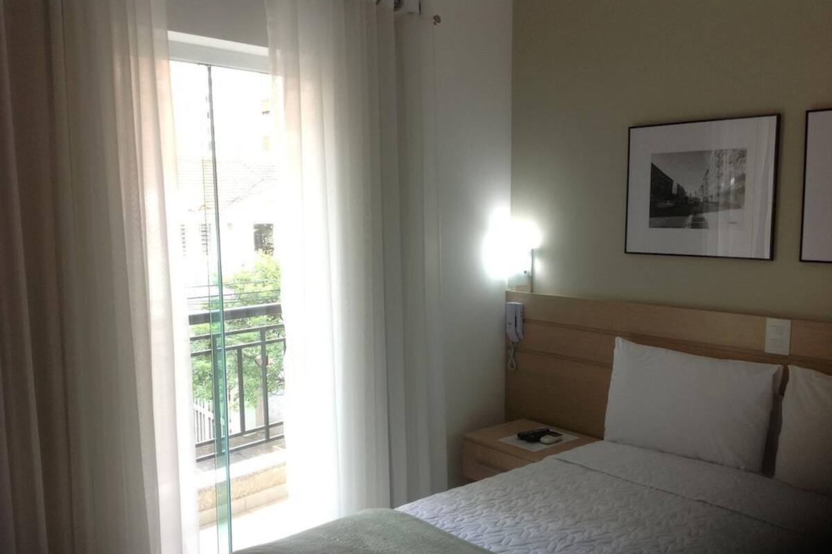 Hotellihuone, jossa on sänky, yöpöytä lampun kanssa ja ikkuna, jossa on verhot.