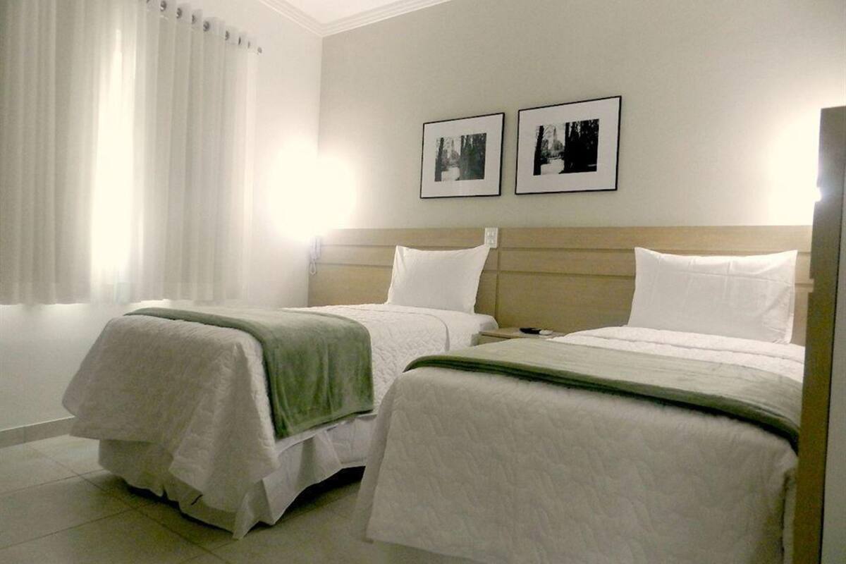 Hotellihuone, jossa on kaksi sänkyä, suuri ikkuna valkoisilla verhoilla ja kaksi kehystettyä kuvaa seinällä.