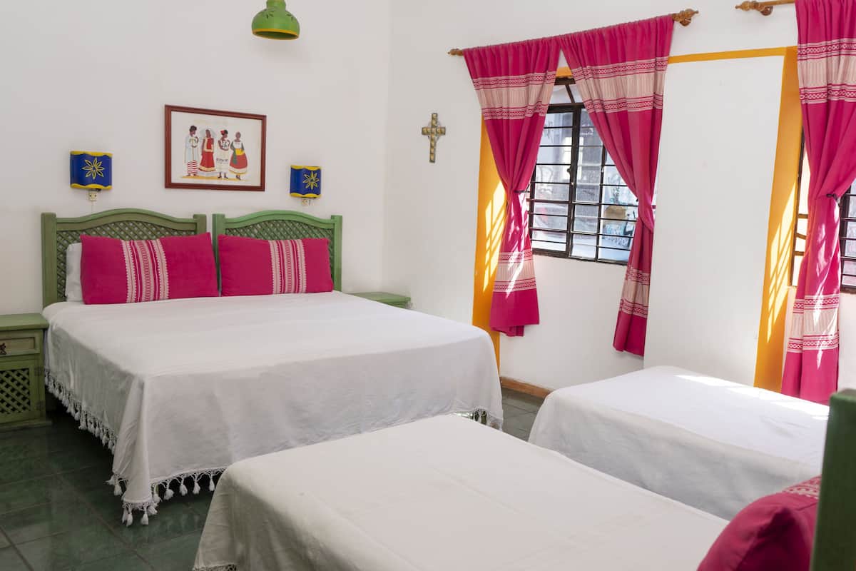 Une chambre avec deux lits, une tête de lit verte, une croix au mur et une fenêtre avec des rideaux roses.