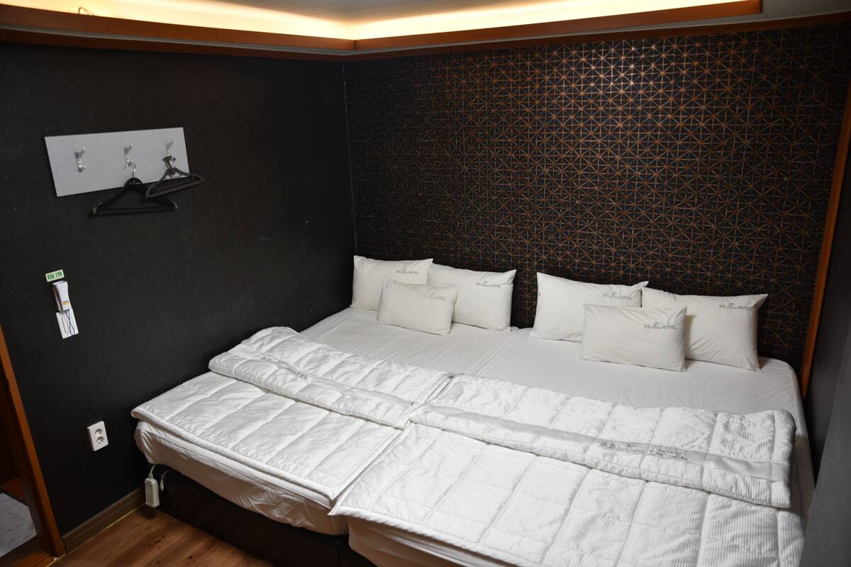 Una cama bien hecha con ropa de cama blanca y varias almohadas, una pared oscura con estampado y una lámpara de pared sencilla.