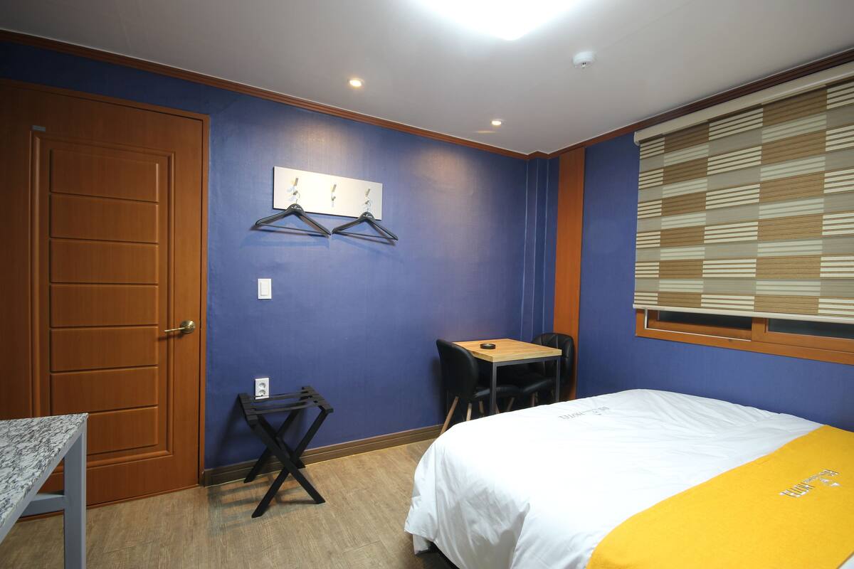 Un dormitorio con cama, escritorio y silla. Se observa una puerta y un ventanal con persianas.