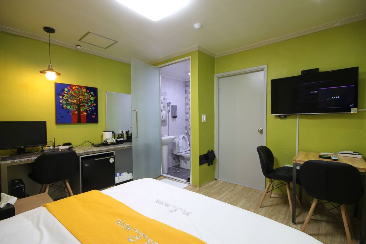 Habitación de hotel con cama, escritorio, sillas, televisión y baño visible a través de una puerta abierta.