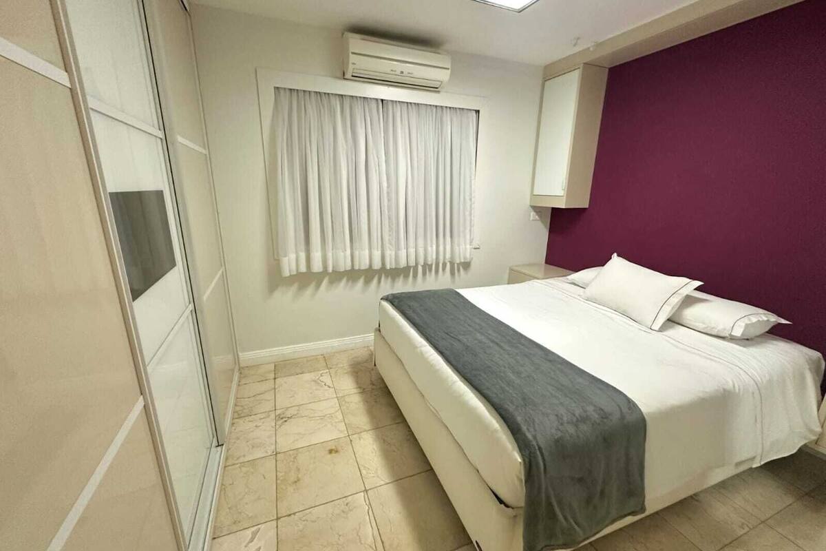 A hotel room with a bed, a nightstand, an air conditioning unit, a window with curtains, and a purple accent wall.