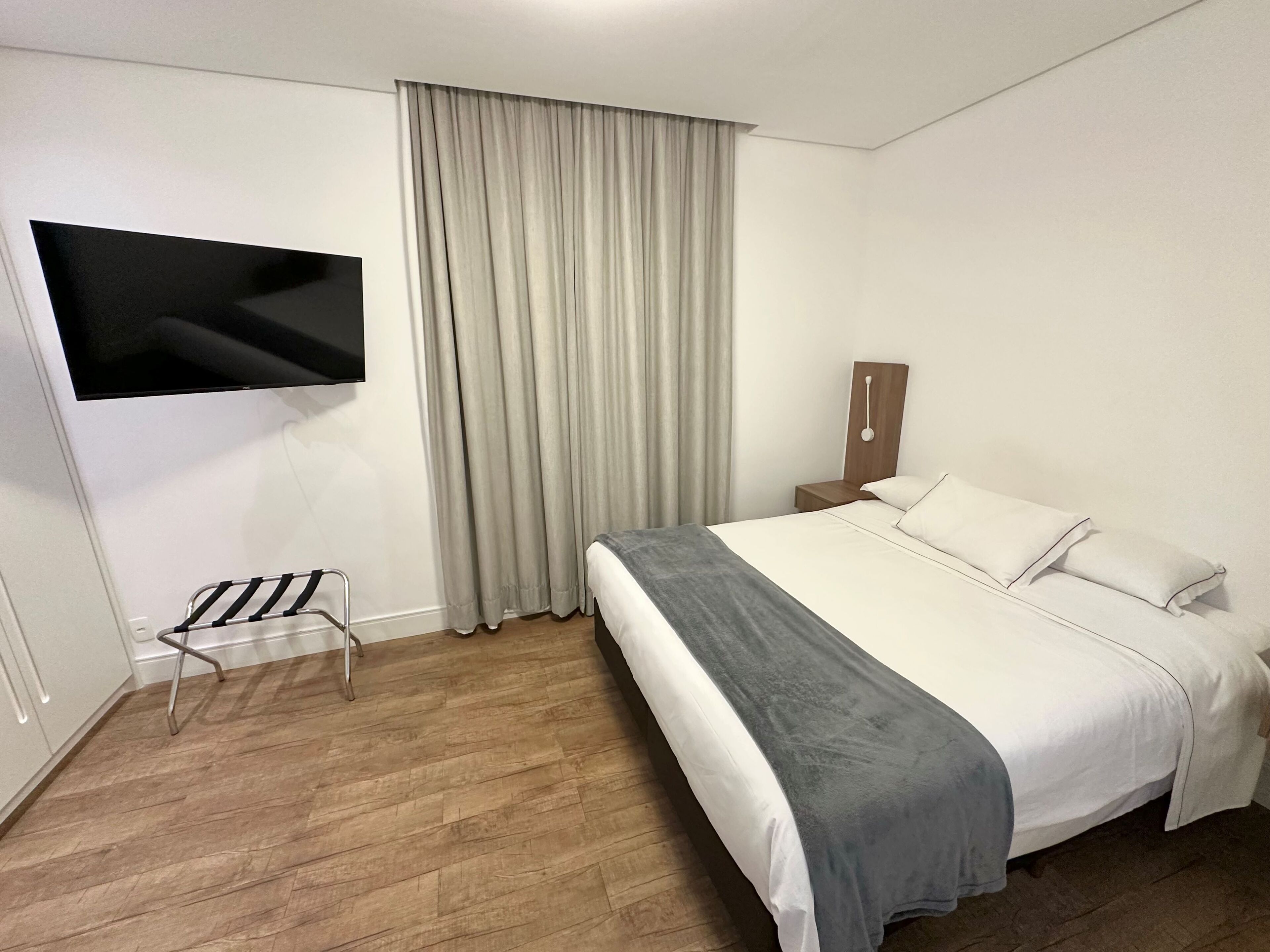 A modern hotel room with a large bed, a flat-screen TV mounted on the wall, a wooden floor, and a window with curtains.