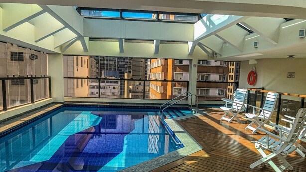 An indoor swimming pool with clear blue tiles, wooden deck, and lounge chairs. - Travel Inn Flats Jardins (São Paulo)