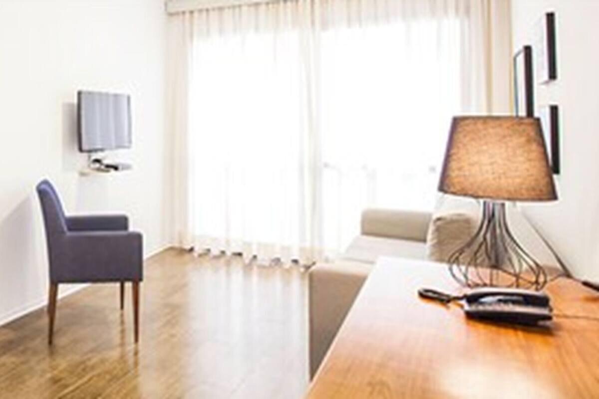 A hotel room with a wooden floor, a desk with a lamp, a chair, a TV mounted on the wall, and a window with curtains.