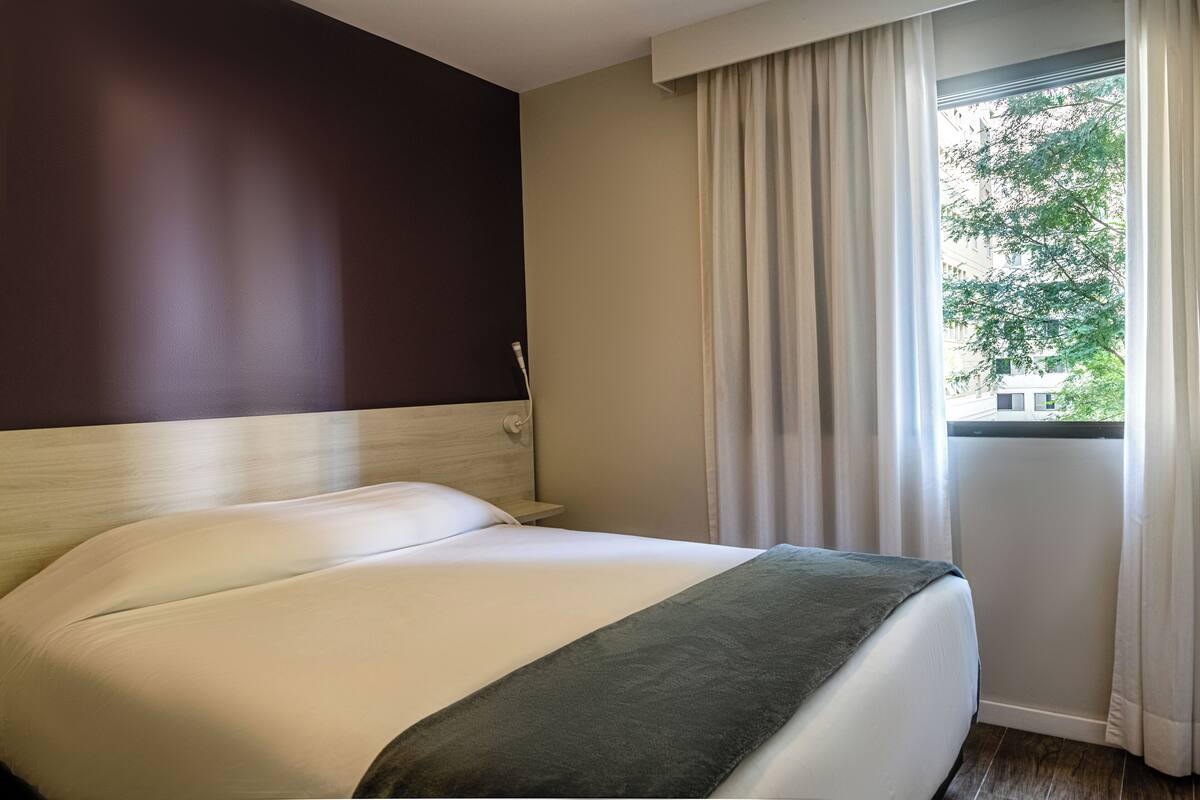 A hotel room with a bed, a nightstand, a window with curtains, and a view of greenery outside.