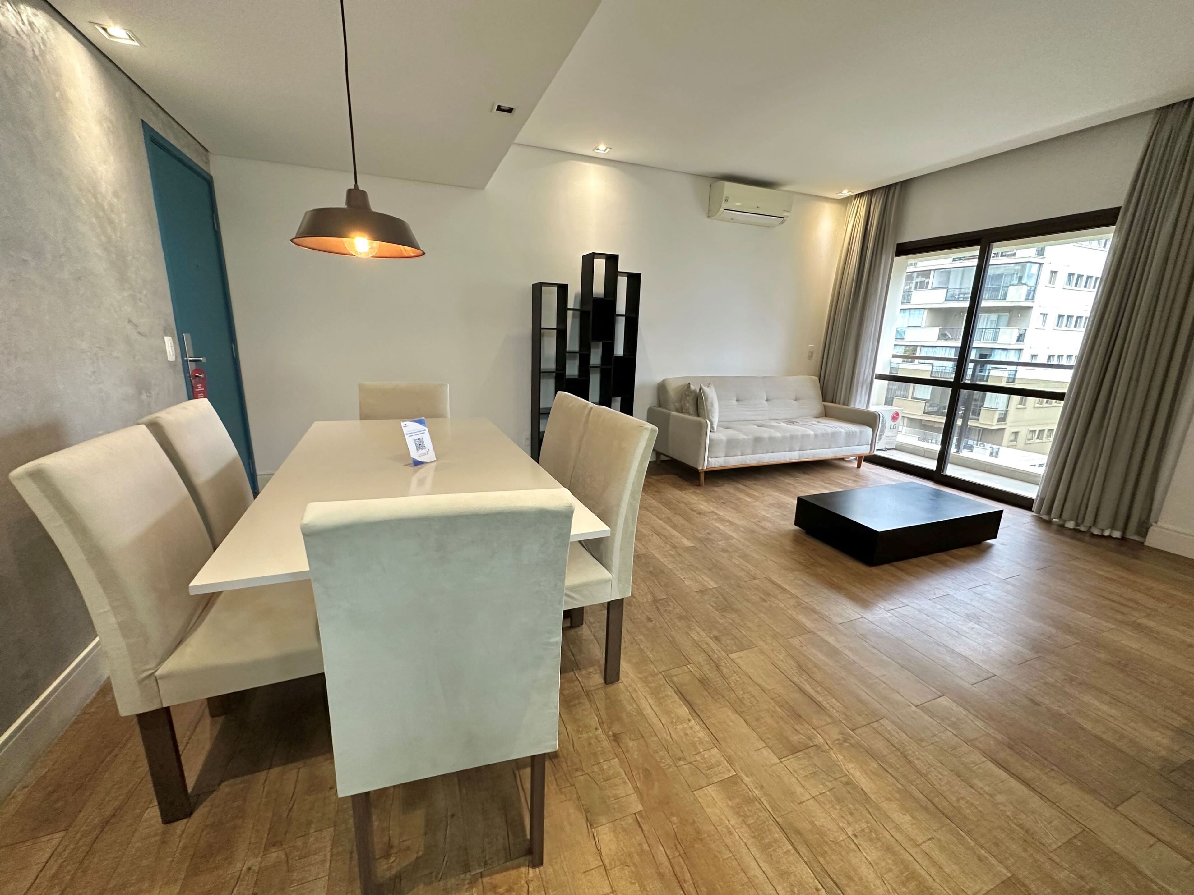 A modern living room with a dining table, chairs, a sofa, a coffee table, and a large window with a view of buildings.