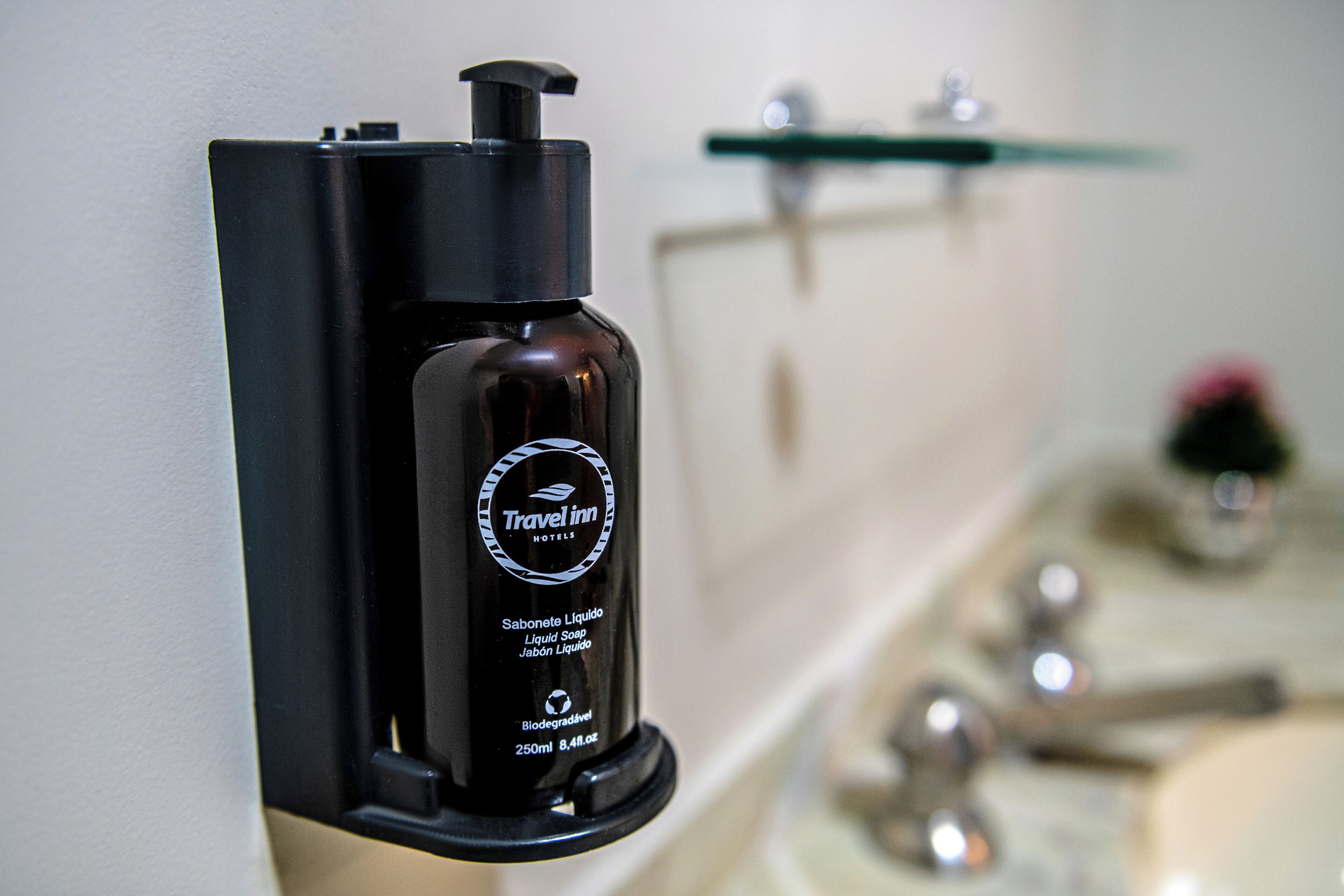 Una botella negra de jabón líquido Travel Inn Hotels con dosificador de bomba, montada en una pared junto a un lavamanos de baño.