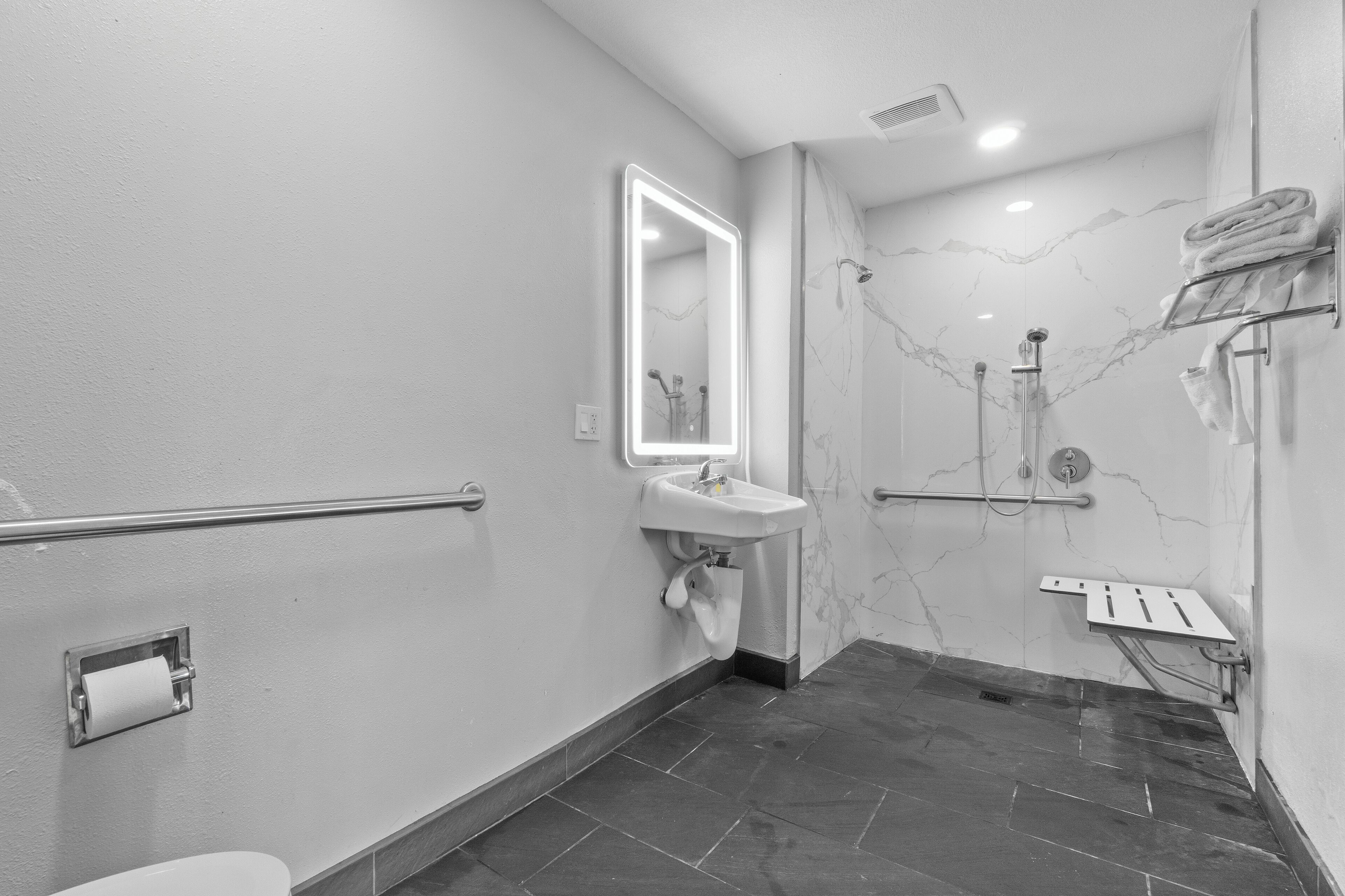 A modern bathroom with a marble wall, a sink with a mirror, a handrail, and a shower area.