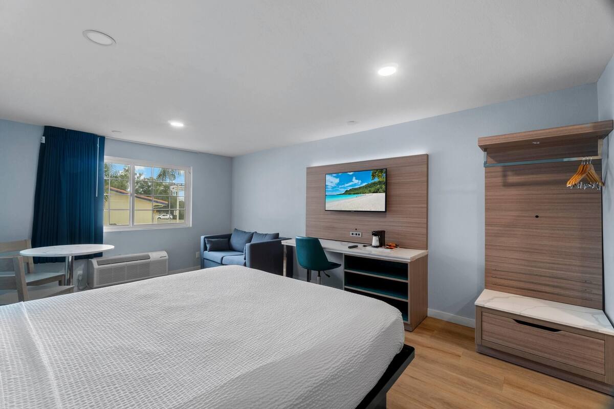 A modern hotel room with a large bed, a desk with a chair, a TV mounted on the wall, and a wardrobe with a hanging rack.
