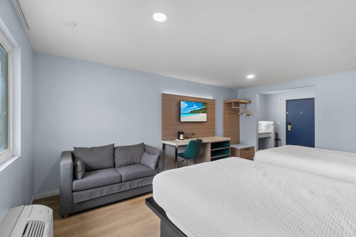 A modern hotel room with a bed, a sofa, a desk with a computer, and a TV mounted on the wall.