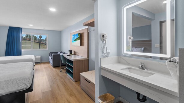 Una habitación de hotel moderna con una cama grande, un escritorio, una silla, una lámpara de escritorio, un espejo amplio, un lavamanos y una ventana con cortinas.
