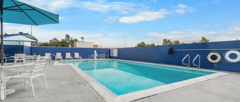 Una piscina con sillones de descanso y sombrillas, rodeada por un muro azul y un cielo despejado.