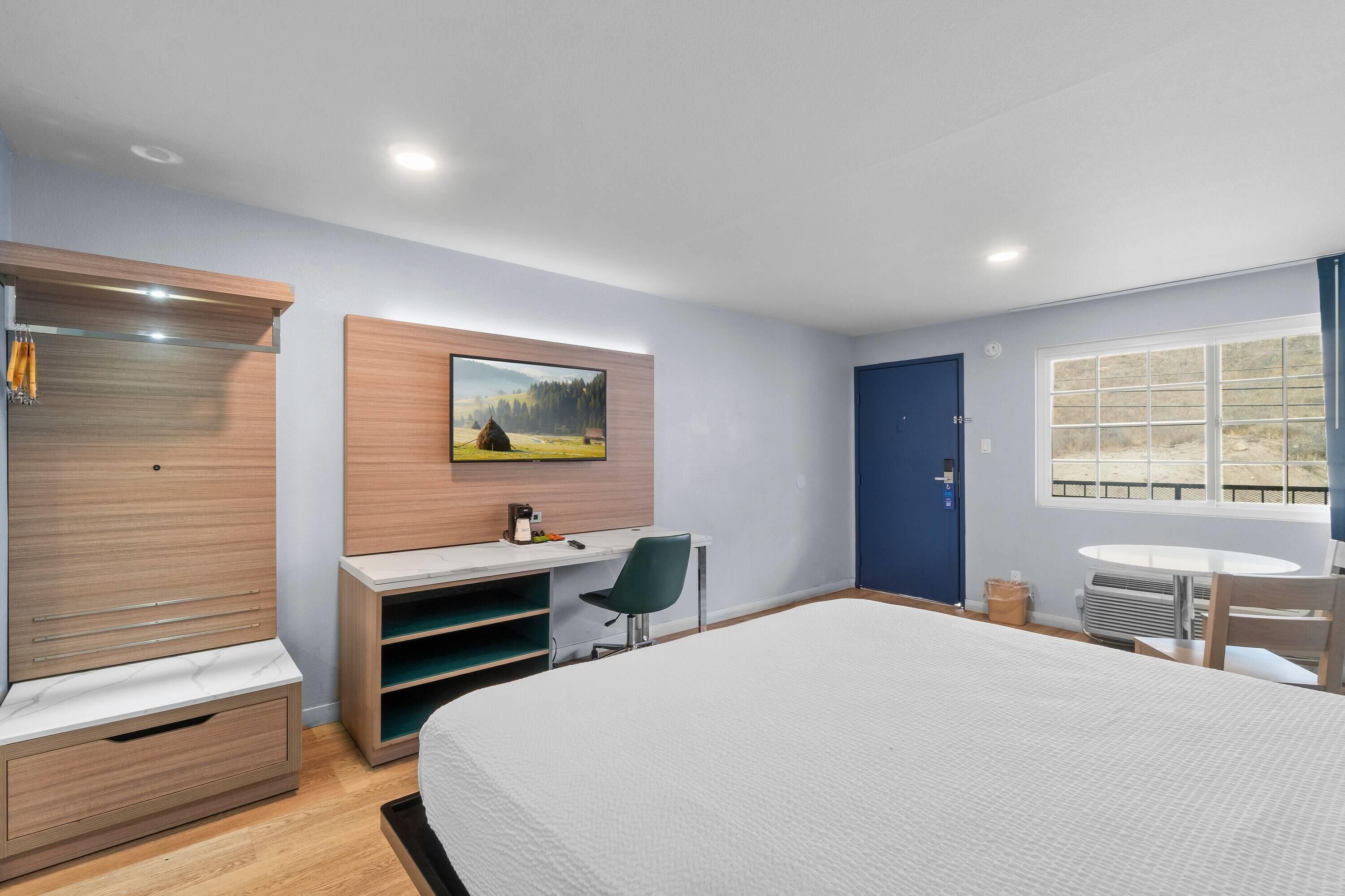 Quarto de hotel com uma cama grande, uma mesa com cadeira, um guarda-roupa, um quadro na parede, uma janela e uma porta.