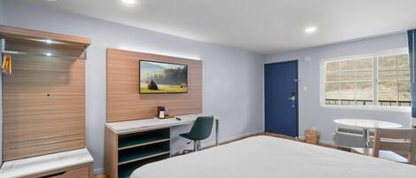 Quarto de hotel com uma cama grande, uma mesa com cadeira, um guarda-roupa, um quadro na parede, uma janela e uma porta.