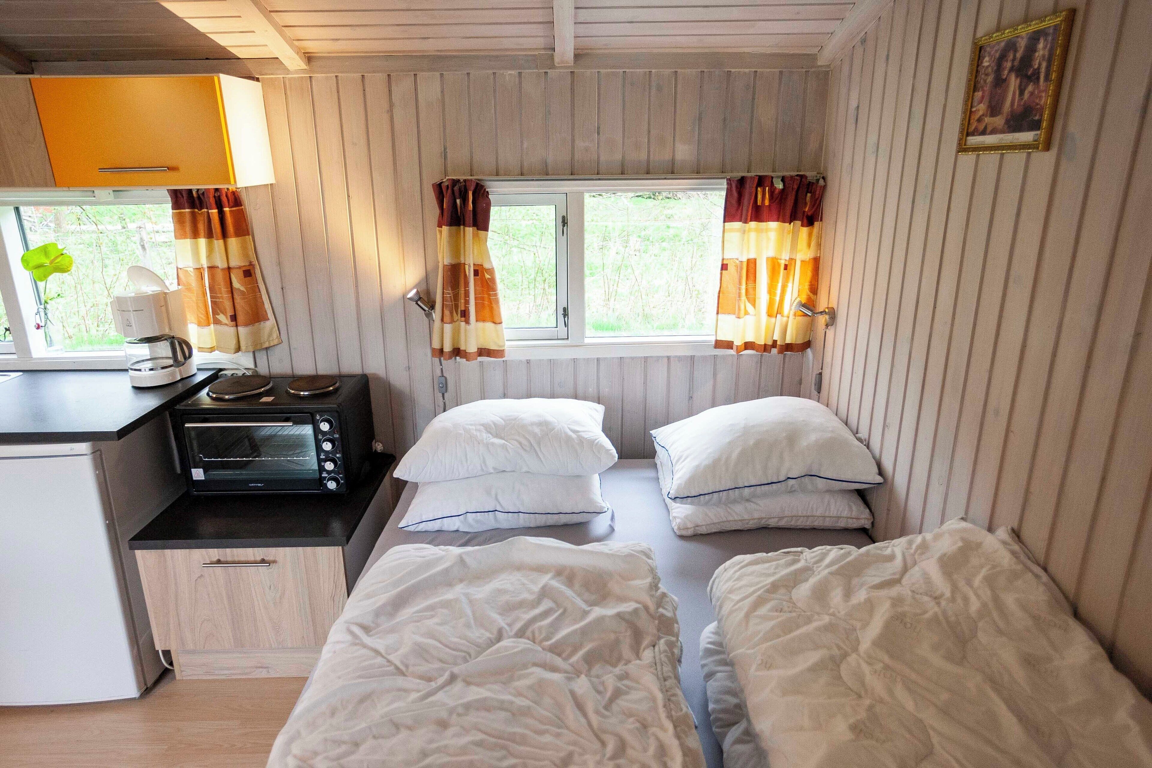Une pièce compacte comprenant un coin cuisine, deux lits, une fenêtre avec des rideaux et une petite table avec une lampe.