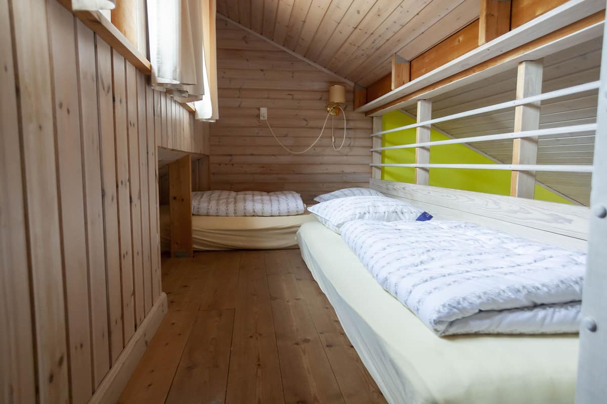 Una pequeña habitación de estilo cabaña con dos camas, una ventana de marco verde y una lámpara fijada a la pared.