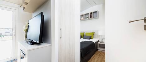 Una habitación de hotel moderna con televisión de pantalla plana, una cama con almohadas, una mesita de noche con lámpara y un pequeño estante con objetos decorativos.