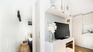 A modern living space with a flat-screen TV on a wooden console, a small table with a lamp, and a vase with white flowers.