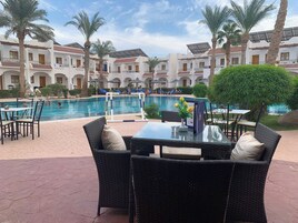 A poolside area with wicker furniture and a glass table, overlooking a swimming pool and a building with multiple balconies.