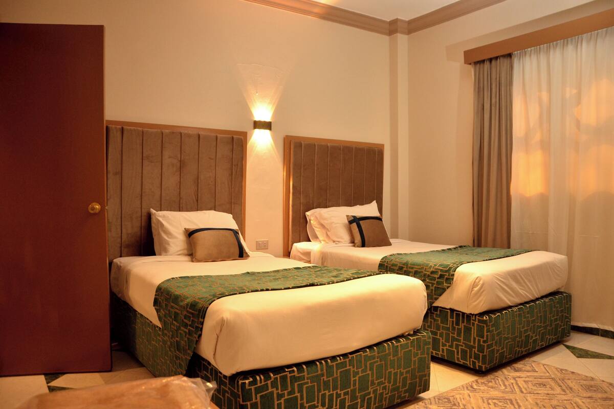 A hotel room with two beds, a wooden headboard, a wall-mounted light, and a window with curtains.