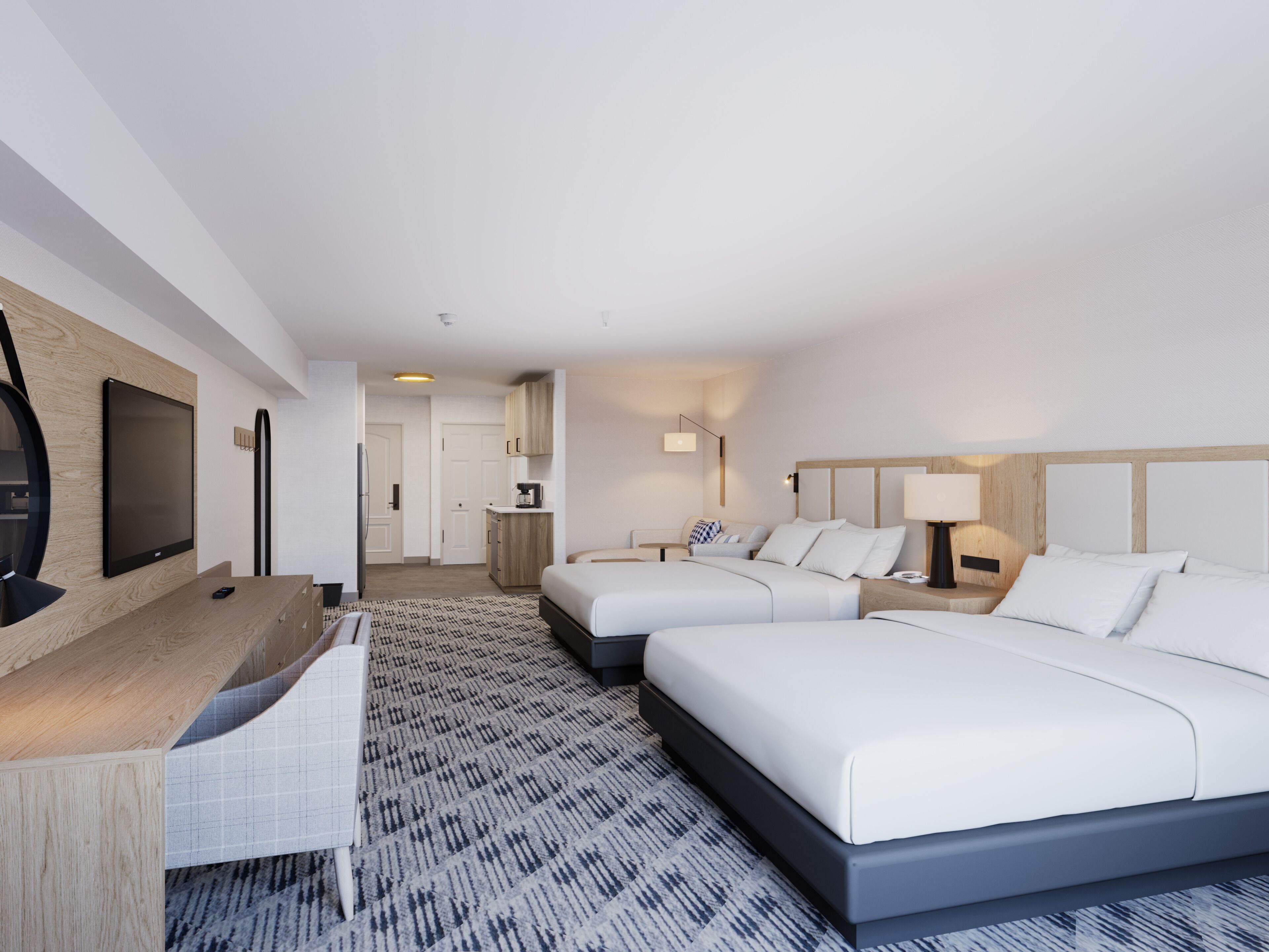 A hotel room with two beds, a desk, a flat-screen TV, a kitchenette, and a patterned carpet.
