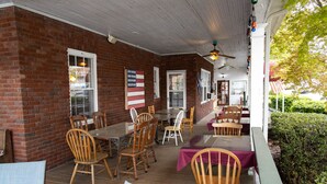 Ein Backsteinhaus mit überdachter Terrasse, auf der Holztische und -stühle stehen, eine amerikanische Flagge weht und die umliegende Grünfläche zu sehen ist.