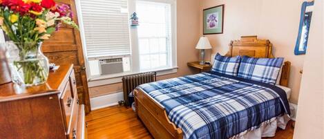 Un dormitorio con cama de madera, cómoda, radiador, ventana con persianas, lámpara y un cuadro en la pared.