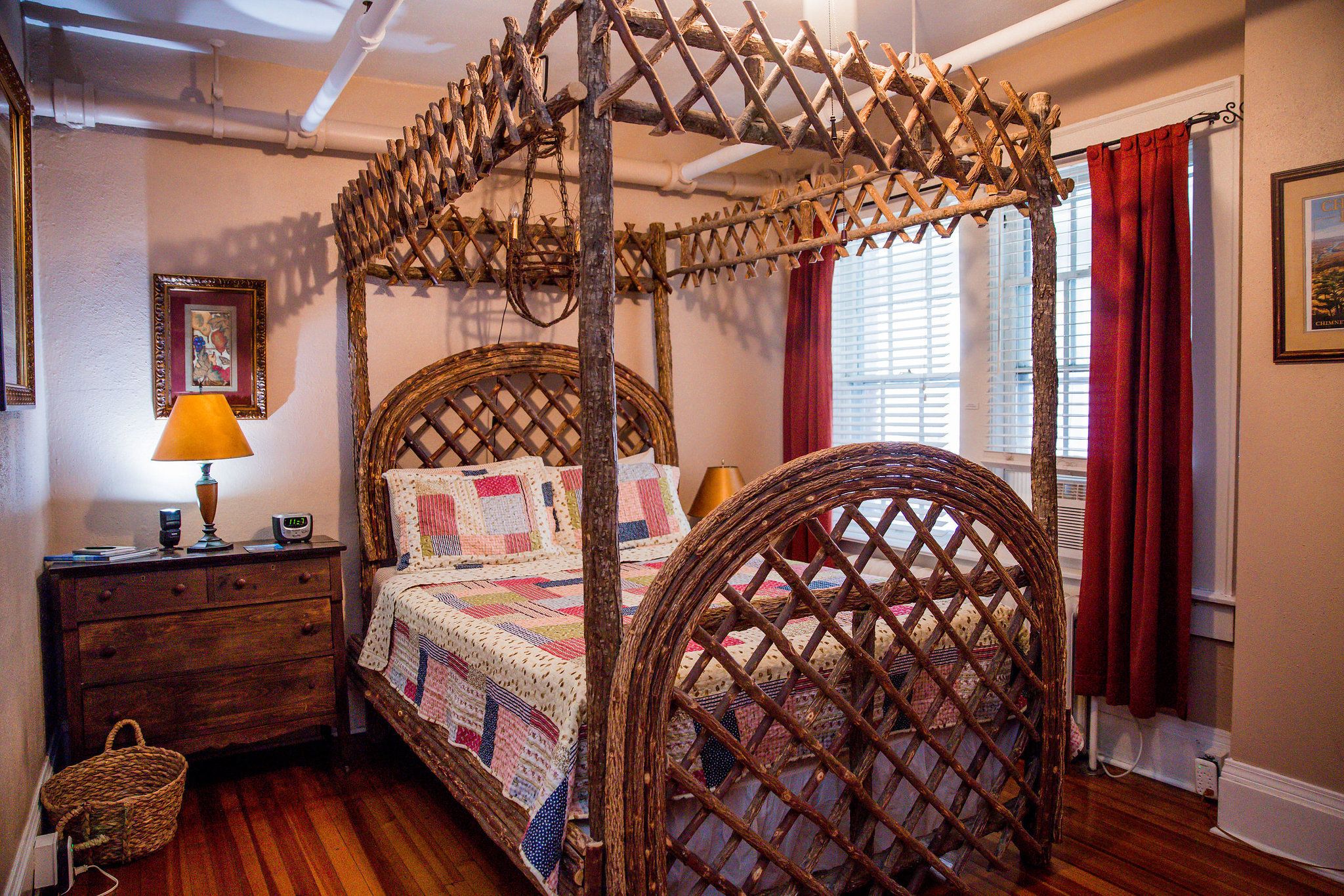 A four-poster canopy bed with a quilt, wooden nightstand, lamp, and wicker chair.