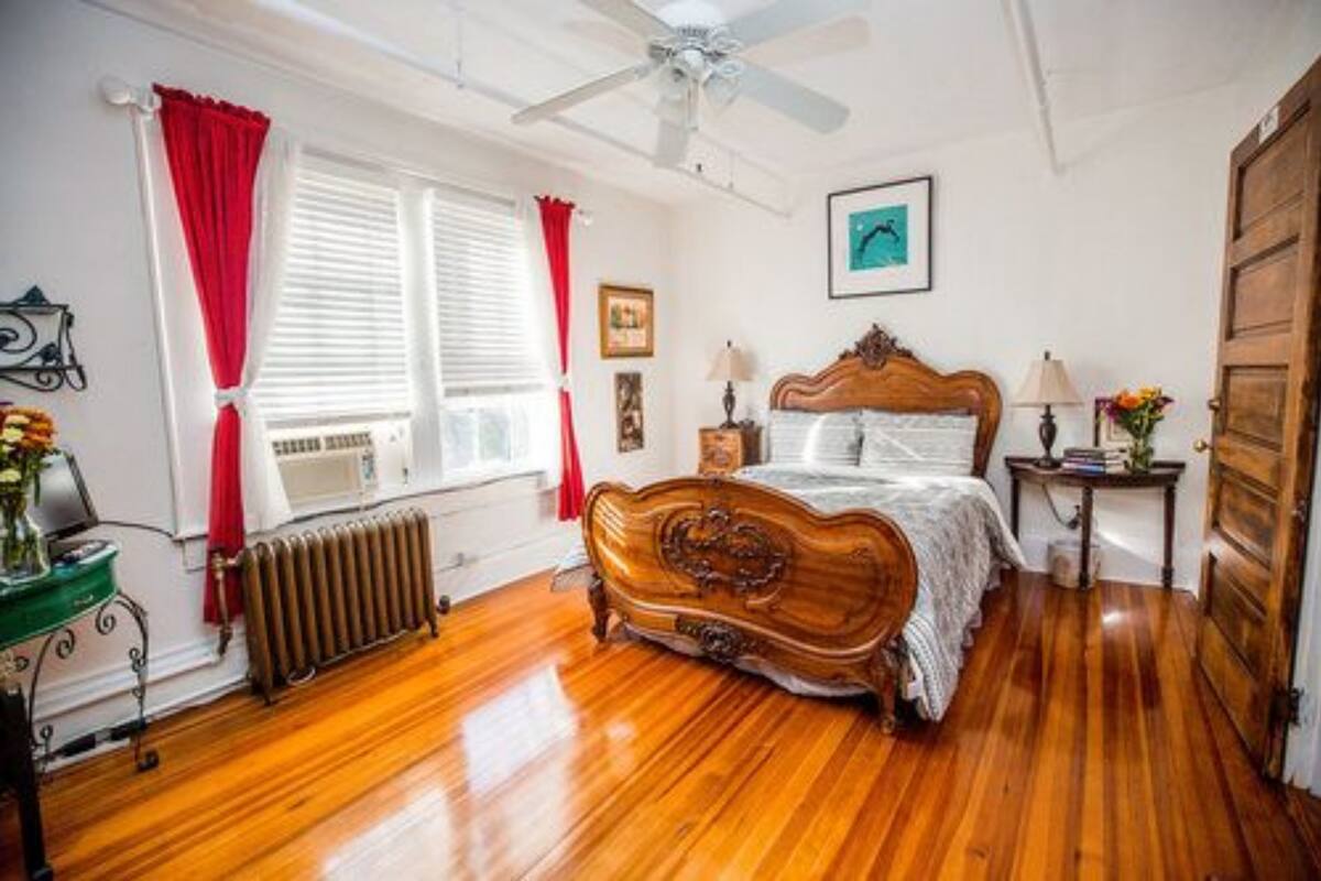 A bedroom with a wooden bed, a radiator, a ceiling fan, and a window with red curtains.