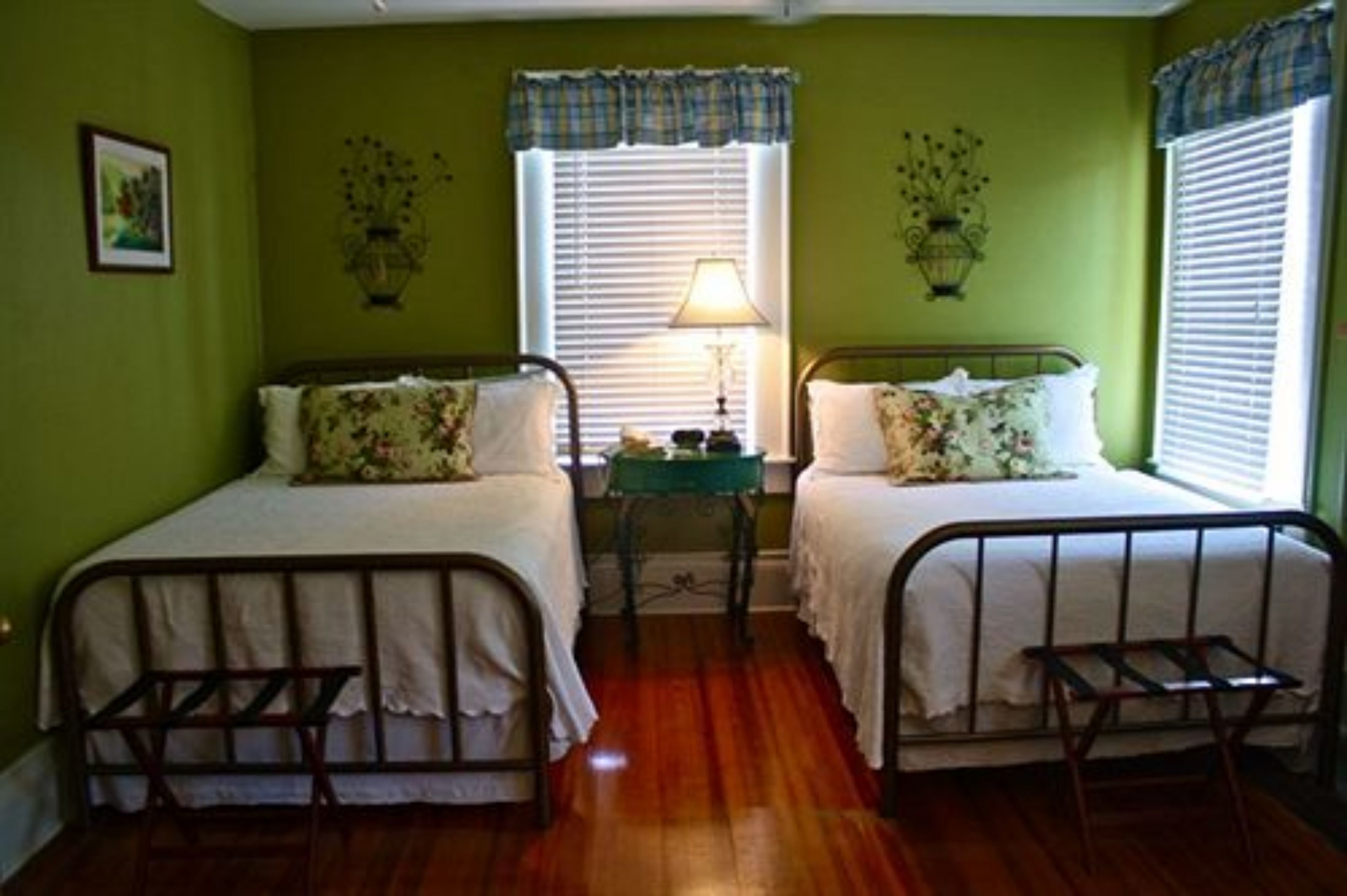A room with two single beds, a small table with a lamp, and a window with blinds.