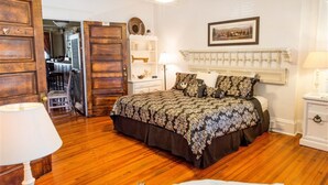 Ein Schlafzimmer mit Holzboden, einem Bett mit gemustertem Überwurf, einem weißen Nachttisch, einer weißen Kommode, einem Deckenventilator und einem gerahmten Bild an der Wand.