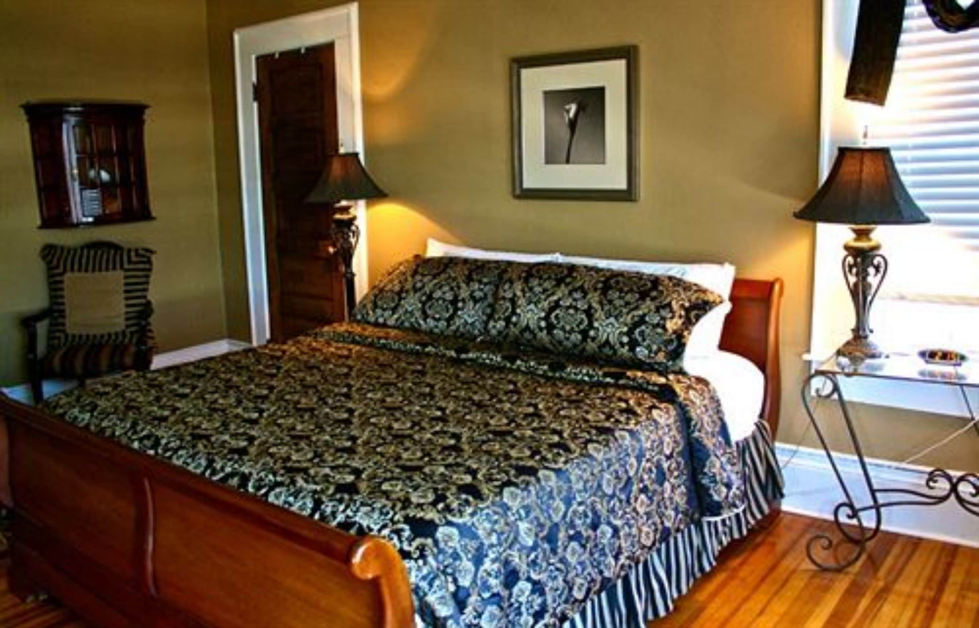 A bedroom with a wooden bed, patterned bedspread, a framed picture on the wall, and a nightstand with a lamp.