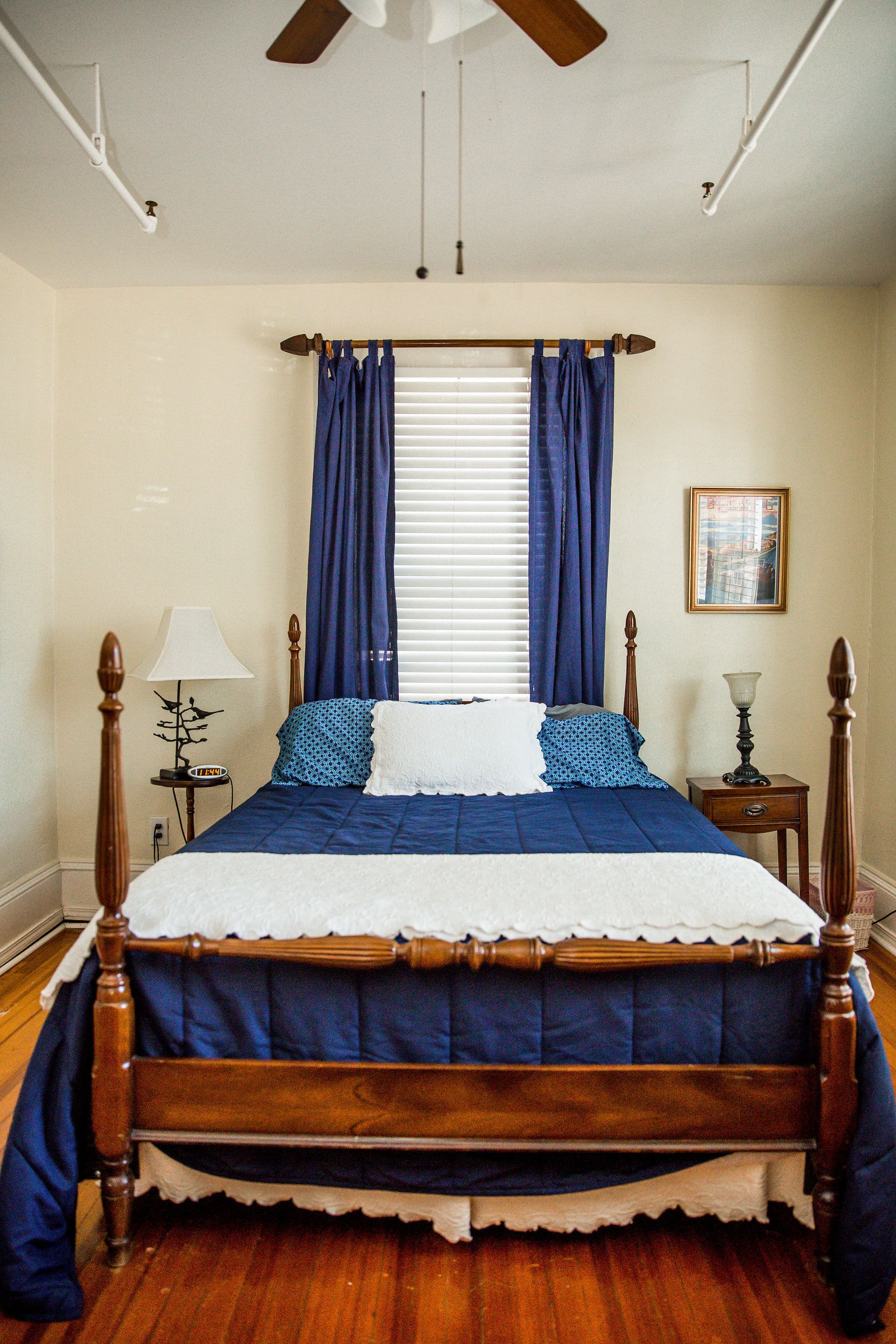 A bedroom with a wooden bed, blue bedding, a nightstand with a lamp, and a ceiling fan.