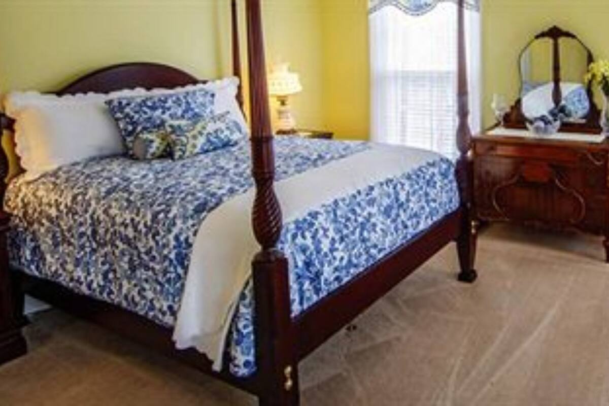 A four-poster bed with a floral bedspread, a wooden nightstand, a dresser with a mirror, and a chair with a floral pattern.