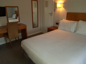 A hotel room with a bed, a desk, a chair, a mirror, a nightstand, and a wall-mounted lamp. - Tri-star Hotel (Birmingham)