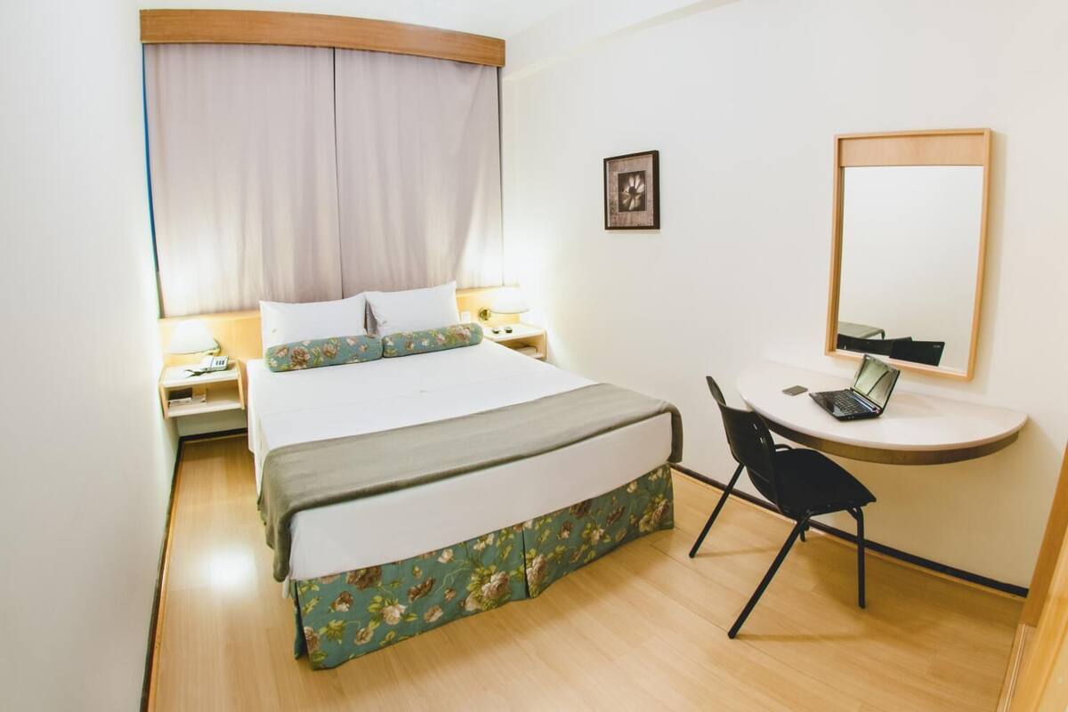 A hotel room with a bed, bedside tables, a desk with a laptop, a chair, a mirror, and a picture on the wall.