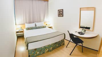 Una habitación de hotel con cama, mesitas de noche, un escritorio con una computadora portátil, una silla, un espejo y un cuadro en la pared.